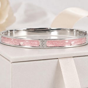 Könnte beinhalten: Ein silberfarbener Klapparmreif mit zwei länglichen, hellrosa glitzernden Harzeinlagen, die durch einen kleinen vertikalen Steg mit drei klaren, rund geschliffenen Kristallen getrennt sind. Der Armreif hat eine polierte Metalloberfläche.