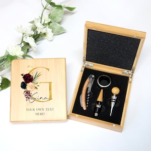 Puede incluir: Una caja de madera con una tapa grabada personalizada que presenta un diseño floral y el nombre "Gina". La caja contiene un sacacorchos, un tapón de botella y un anillo de extracción.