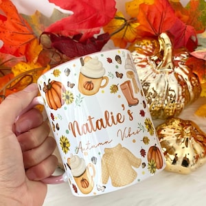 Puede incluir: Taza de cer&aacute;mica blanca con asa rosa. La taza tiene un dise&ntilde;o de oto&ntilde;o con calabazas, hojas, un su&eacute;ter y una taza de caf&eacute;. El texto "Natalie's Autumn Vibes" est&aacute; impreso en la taza.