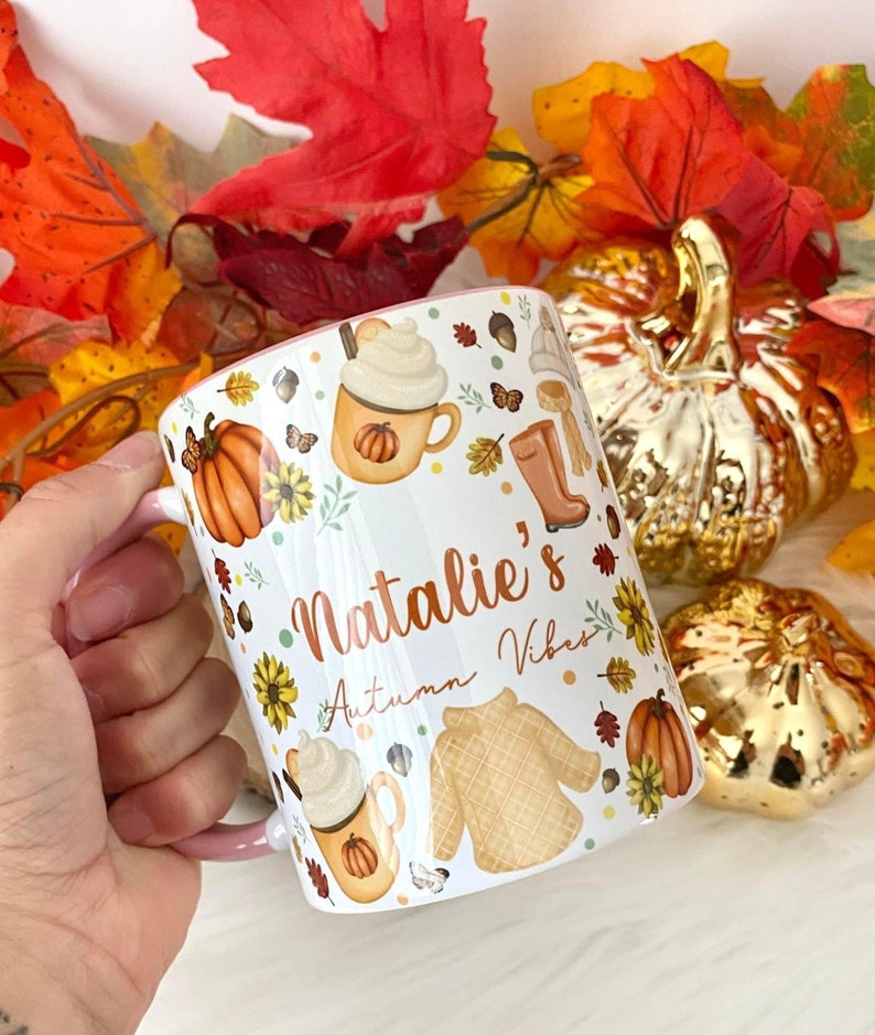 Puede incluir: Taza de cer&aacute;mica blanca con asa rosa. La taza tiene un dise&ntilde;o de oto&ntilde;o con calabazas, hojas, un su&eacute;ter y una taza de caf&eacute;. El texto "Natalie's Autumn Vibes" est&aacute; impreso en la taza.