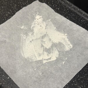 May include: A square of parchment paper with a white, textured substance spread across it. The paper sits on a dark, speckled surface, possibly a baking sheet or tray. The substance appears to be a paste or powder.