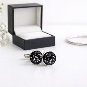 Puede incluir: Un par de gemelos plateados con un diseño moteado negro y plateado. Los gemelos se muestran junto a una caja de joyería negra abierta y una pulsera plateada. Flores blancas en el fondo.