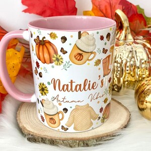 Puede incluir: Taza de cer&aacute;mica blanca con borde y asa de color rosa. La taza presenta un dise&ntilde;o de tem&aacute;tica oto&ntilde;al con calabazas, hojas, mariposas y un su&eacute;ter. El texto "Natalie's Autumn Vibes" est&aacute; escrito en la taza.