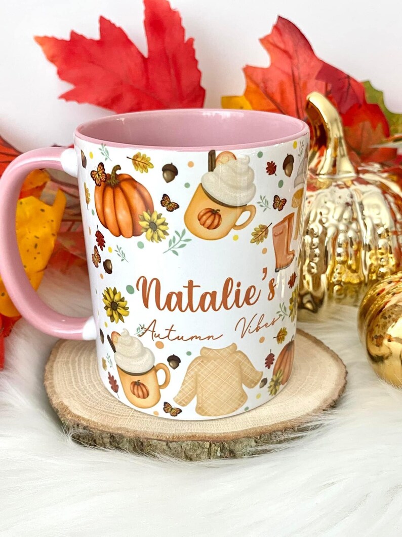 Puede incluir: Taza de cer&aacute;mica blanca con borde y asa de color rosa. La taza presenta un dise&ntilde;o de tem&aacute;tica oto&ntilde;al con calabazas, hojas, mariposas y un su&eacute;ter. El texto "Natalie's Autumn Vibes" est&aacute; escrito en la taza.