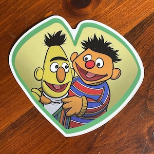 May include: A sticker featuring Bert and Ernie from Sesame Street. Bert is yellow with a white shirt and Ernie is orange with a red nose and a striped shirt. The sticker has a green border and a yellow background.