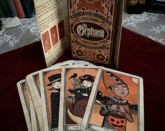 May include: A set of tarot cards with illustrations of children in various costumes. The cards are laid out on a red surface with a box that reads "The Orphans Divining the Future" in gold lettering. The box is decorated with an eye and ornate details.