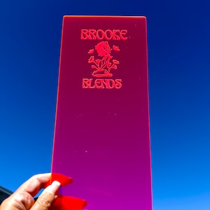 May include: A rectangular, neon pink acrylic sign with the text "BROOKE BLENDS" in red. The sign features a floral design in the center. The sign is held up against a bright blue sky.