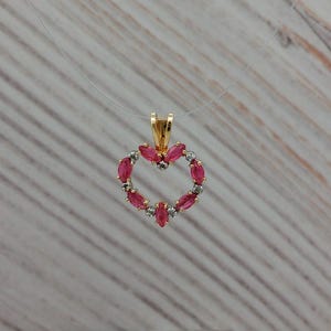 May include: A gold pendant necklace featuring a heart-shaped design. The heart is outlined with pink gemstones and interspersed with small, clear stones. The pendant hangs from a thin, clear chain.