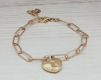 May include: Gold-colored bracelet featuring a heart-shaped padlock charm. The bracelet has a textured chain link design and a lobster clasp closure. The heart charm has a keyhole detail. The bracelet is displayed on a light-colored wooden surface.
