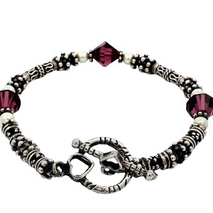 May include: A silver-toned bracelet featuring a toggle clasp. The bracelet is adorned with dark purple faceted beads, small white beads, and detailed silver-toned metal beads. The design is symmetrical, with the clasp at the bottom.