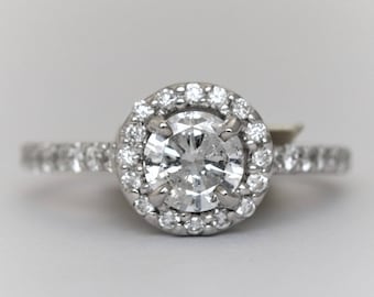 Könnte beinhalten: Ein silberner Verlobungsring mit einem runden, klaren Diamanten in der Mitte, der von einem Heiligenschein aus kleineren Diamanten umgeben ist. Die Ringschiene ist ebenfalls mit kleineren Diamanten besetzt.
