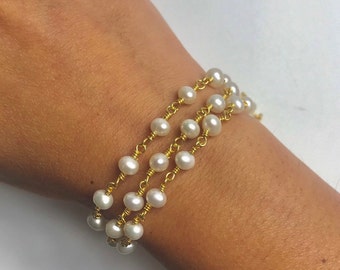 Puede incluir: Un brazalete de cadena dorada con perlas blancas. El brazalete está formado por tres hileras de perlas.