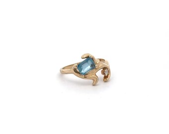 May include: A gold ring with a blue gemstone and a smaller clear gemstone. The ring has a unique, organic design.