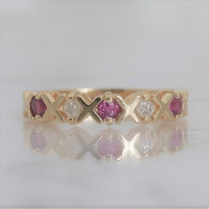 Puede incluir: Un anillo de banda de oro con piedras preciosas rosas y blancas alternadas. La banda está decorada con un patrón de X repetido.