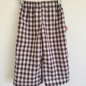 Puede incluir: Shorts a cuadros marrones y blancos colgados de una percha de madera. Los shorts tienen una cintura elástica y una pequeña etiqueta roja en el lateral. La tela presenta un clásico estampado de cuadros vichy.