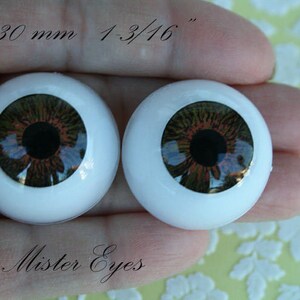 May include: Two brown and black doll eyes with a white iris and a black pupil. The eyes are 30 mm or 1-3/16 inches in diameter. The eyes are on a hand with the text "Mister Eyes" below them.
