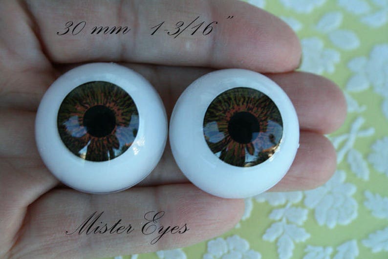 May include: Two brown and black doll eyes with a white iris and a black pupil. The eyes are 30 mm or 1-3/16 inches in diameter. The eyes are on a hand with the text "Mister Eyes" below them.