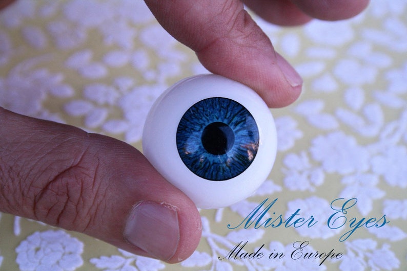 May include: A close-up of a blue glass eye with a white sclera. The eye is being held by a hand. The text "Mister Eyes Made in Europe" is visible below the eye.