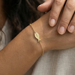 Peut inclure: Un bracelet doré avec un pendentif en forme de poisson. Le pendentif porte le nom "ALICE". Le bracelet est porté au poignet.