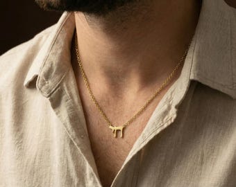 May include: A gold-toned metal necklace featuring a pendant shaped like the Hebrew letter Chai. The pendant is suspended from a delicate, matching gold-toned cable chain. The necklace rests against a light beige, textured linen-style collared shirt.