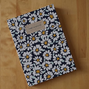 Peut inclure: Un carnet rectangulaire avec un motif floral bleu marine représentant des marguerites blanches à cœur jaune. Le carnet porte une étiquette rectangulaire avec l'inscription "CARNET DE RECETTES".
