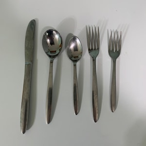 Puede incluir: Un juego de cinco cubiertos plateados, que incluye un cuchillo, dos cucharas y dos tenedores, dispuestos sobre una superficie blanca.