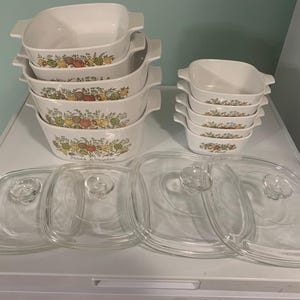 Op de afbeelding: Een set witte keramische ovenschaaltjes met bloemmotieven en deksels. De schaaltjes zijn op elkaar gestapeld en staan op een wit oppervlak. Er zijn ook vier heldere glazen deksels met handvatten.