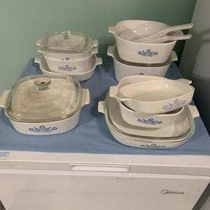 Op de afbeelding: Een set witte ovenschaal met blauwe bloemenmotieven en glazen deksels. De schalen zijn gestapeld op een blauw oppervlak.