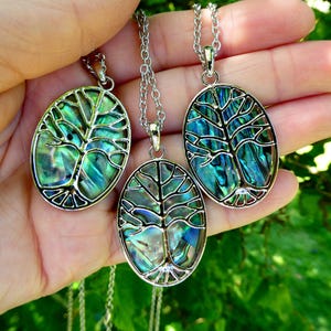 May include: Three oval pendants with silver chains. Each pendant has a tree design with a colourful, iridescent abalone shell inlay. The pendants are held in a hand against a blurred green background.