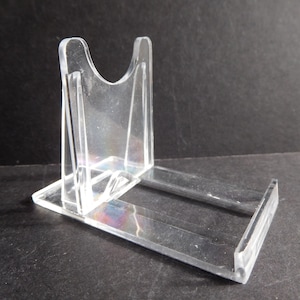 May include: A clear plastic display stand for a small object, such as a collectible or a figurine.