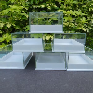 May include: A set of six clear glass display boxes with white bases stacked in a pyramid shape. The boxes are empty and ready to be filled with small items.
