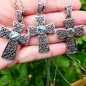 May include: Three silver cross pendants with detailed scrollwork and abalone shell inlays. Each cross is suspended from a fine chain. The pendants are displayed against a background of green leaves.