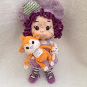 May include: A handmade crochet doll with purple hair and a matching hat, holding an orange and white crochet monkey. The doll wears a purple dress, white shirt, and purple shoes. The doll is approximately 8 inches tall.