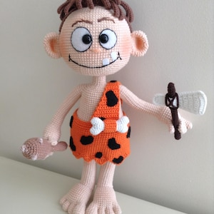 May include: A crocheted plush toy of a caveman with brown hair, wearing an orange and black loincloth, holding a brown club in one hand and a white axe in the other.