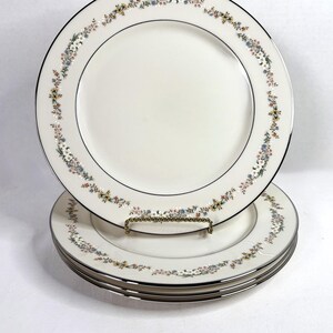 May include: Set of white ceramic plates with a floral border in pastel colors. Each plate has a silver rim and a black inner line. The plates are stacked on a gold-colored metal stand, showcasing a classic dinnerware design.