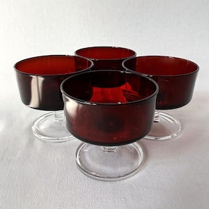 May include: Four vintage ruby red glass dessert bowls. The bowls have a rounded shape with a clear, circular base. The glass has a rich, dark red color, and the bowls are arranged on a white surface.