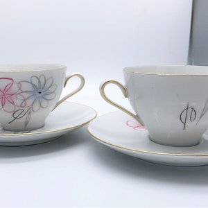 May include: Two white teacups and saucers with gold trim. One cup features pink and blue floral designs, while the other has a minimalist black and gray design. The cups have gold handles and are set on matching saucers.
