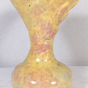 May include: A ceramic vase with a flared top and a bulbous base. The vase is primarily yellow with pink and peach-colored accents. The design includes a sculpted floral element on the side. The vase has a glossy finish.
