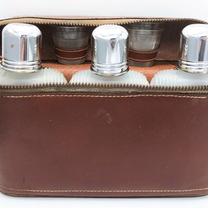 May include: A brown leather travel bar set with a zippered closure. The set includes three frosted glass flasks with silver caps and two shot glasses. The shot glasses are wrapped with brown leather bands.