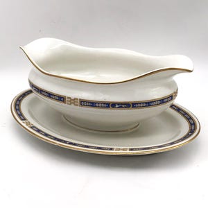 May include: White porcelain gravy boat with a matching oval-shaped underplate. The gravy boat and plate are trimmed with gold and feature a blue and gold decorative band. The gravy boat has a curved spout.