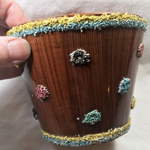 Puede incluir: Una maceta de cer&aacute;mica marr&oacute;n con acabado brillante y textura de vetas de madera verticales. El borde y la base est&aacute;n decorados con chispas amarillas, azules y blancas. Peque&ntilde;os grupos coloridos de chispas est&aacute;n dispersos por la superficie de la maceta.