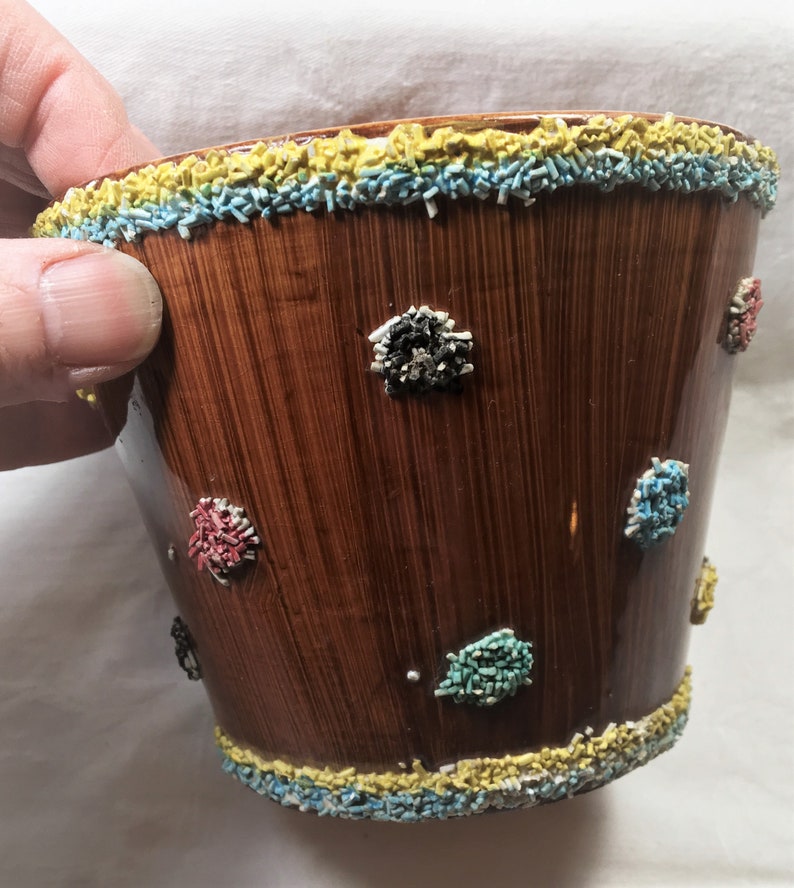 Puede incluir: Una maceta de cer&aacute;mica marr&oacute;n con acabado brillante y textura de vetas de madera verticales. El borde y la base est&aacute;n decorados con chispas amarillas, azules y blancas. Peque&ntilde;os grupos coloridos de chispas est&aacute;n dispersos por la superficie de la maceta.