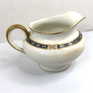 May include: A white ceramic creamer with a gold handle and trim. The creamer features a blue and gold band around the body with a decorative pattern. The creamer is approximately 4 inches tall.