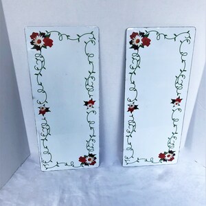 May include: Two white rectangular decorative panels with a floral design. Each panel features a border of green vines and red and white flowers. The panels are slightly angled, showcasing their design and shape.