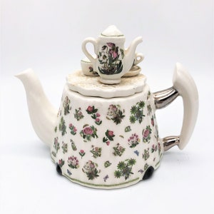 May include: A white ceramic teapot with a floral pattern of pink, green, and purple flowers. A miniature teapot and teacups sit on the lid. The handle and spout are white, with silver accents. The teapot has a curved spout and a large handle.