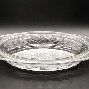May include: Clear glass oval baking dish with floral embossed design. The dish has a rim with handles and a decorative floral pattern around the edge. The bottom of the dish also has a floral design.