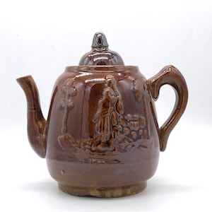 May include: Brown ceramic teapot with a lid and handle. The teapot features a relief design of a person and foliage. The spout and handle are curved. The teapot is glazed with a glossy finish.