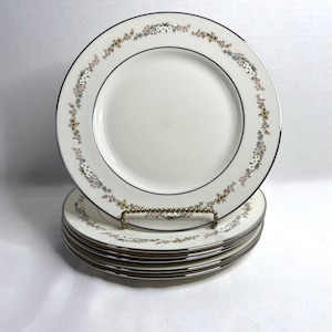 May include: A set of white ceramic plates with a delicate floral border in pastel colors. Each plate has a black and silver trim. The plates are stacked on a gold-colored metal plate stand.