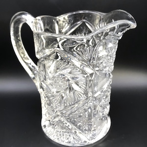 May include: Clear glass pitcher with a handle and a decorative, cut-glass pattern. The pitcher has a scalloped rim and a rounded base. The cut-glass design features geometric shapes and starburst patterns, creating a sparkling effect. The pitcher is likely used for serving beverages.