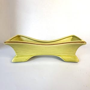 May include: A light green ceramic planter with a curved design. The planter has a textured, ribbed surface and a gold-colored trim along the top edge. It features a unique, arched shape and is supported by two flared feet.